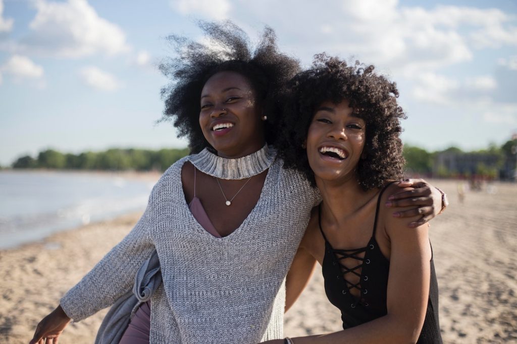 two girls smile on the beach with their arms around each other's shoulders