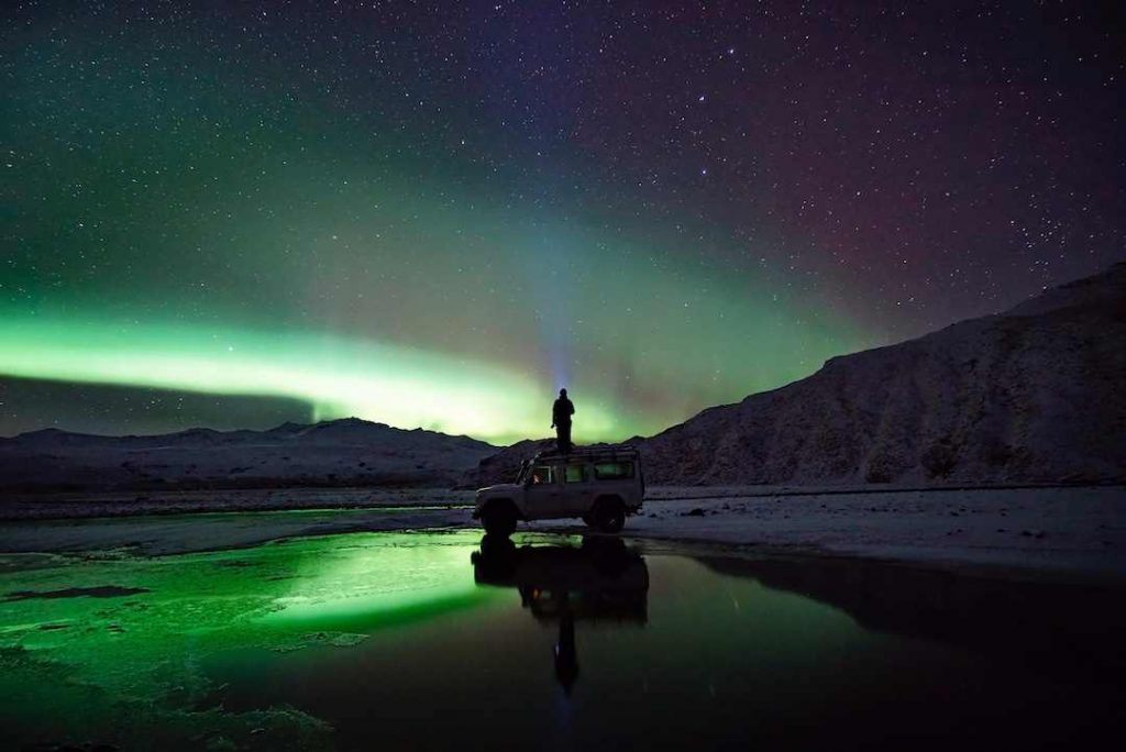 a man standing on top of the roof of a car watchin iceland's northern lights