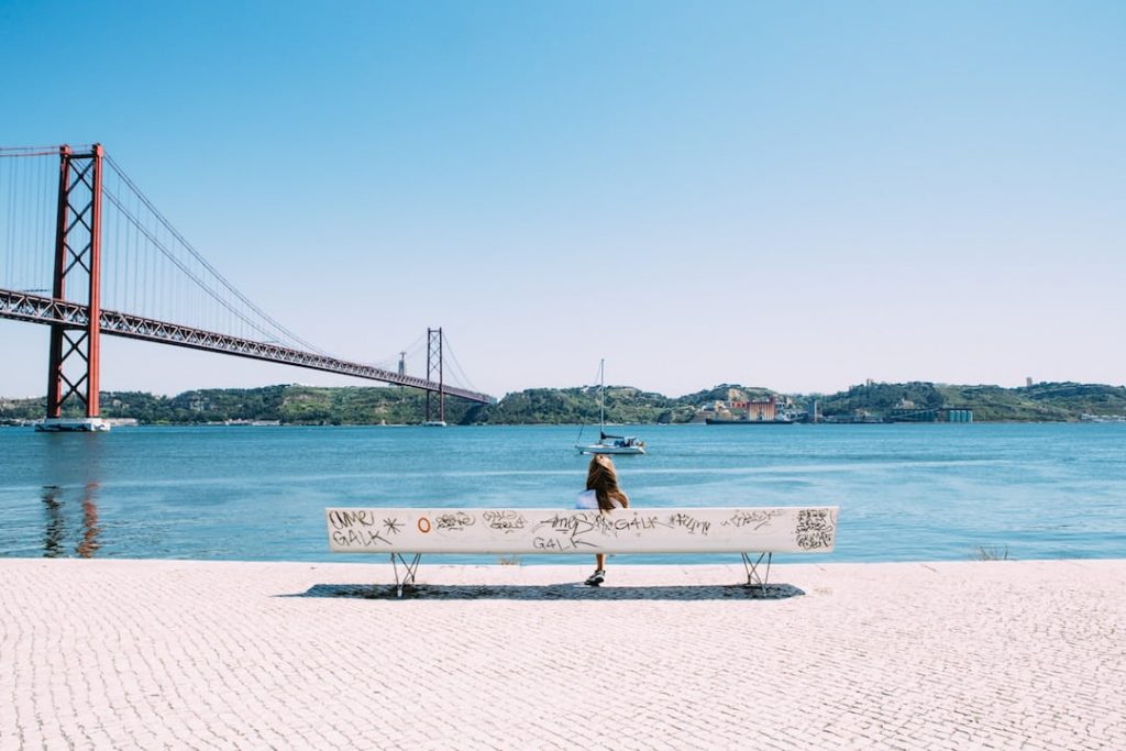 A girl sits on a bench at lookout point by looking out 25th April Bridge, Lisbon, Portugal