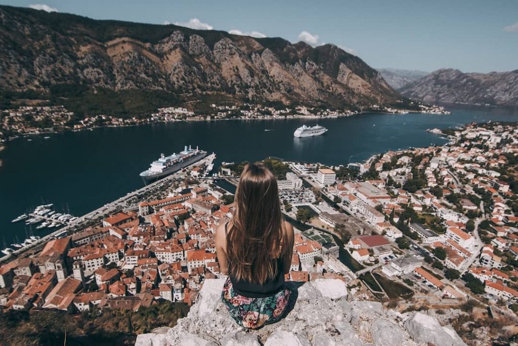 A girl sits with her back to us on the edge of a rock overlooking the bay of Kotor