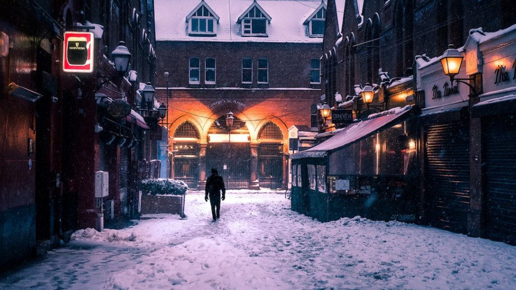 A man walks through a snowy Dublin street, with cosy lighting and pubs