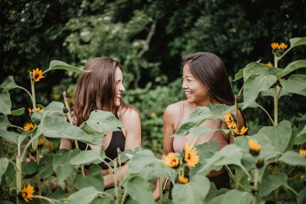 Two girls sit laughing together in nature surrounded by sunflowers