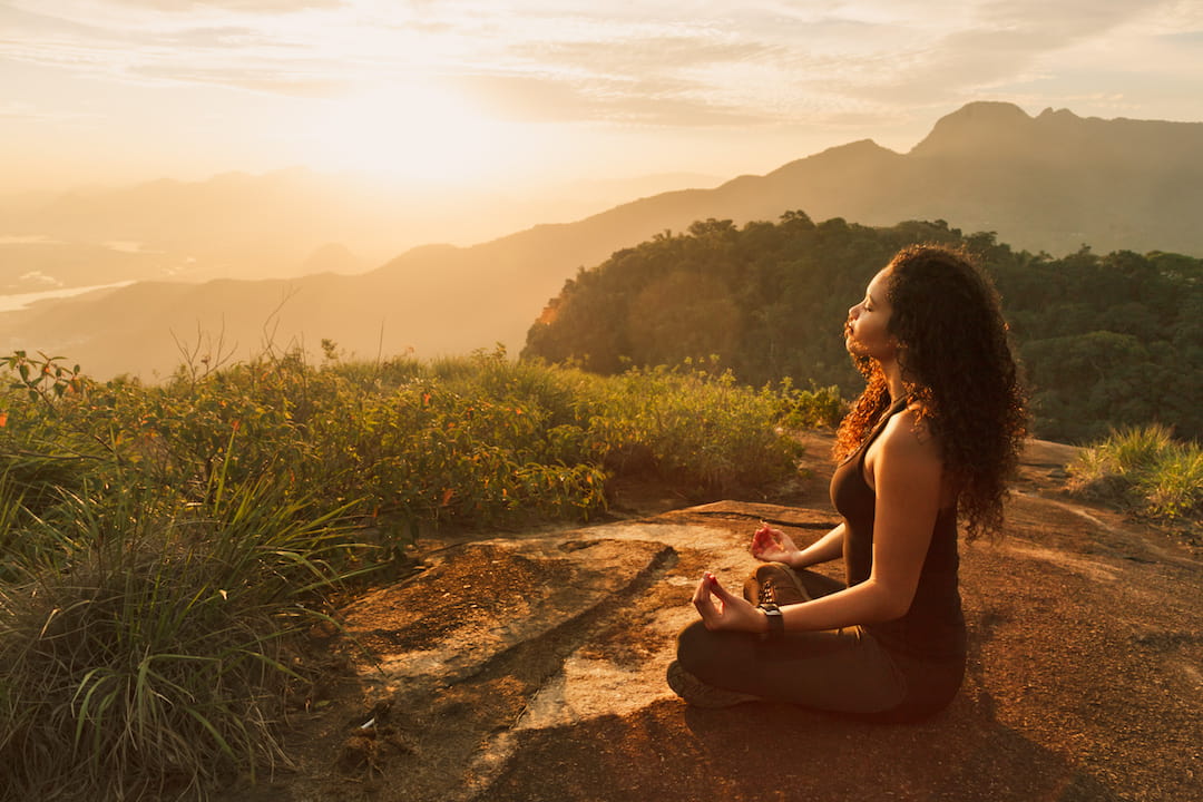 A woman sits cross-legged on a mountain at sunrise, facing the sun with her eyes closed