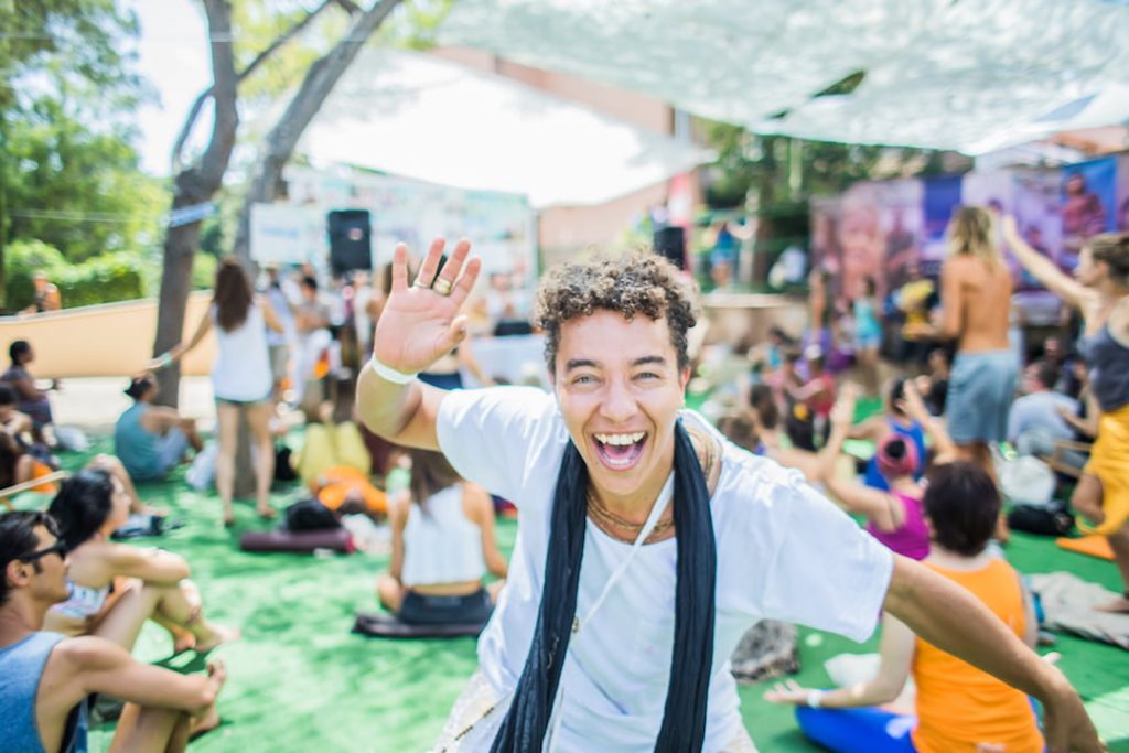 a person in a white shirt and scarf smiles and waves at the camera in an outdoor setting at the Barcelona Yoga Conference