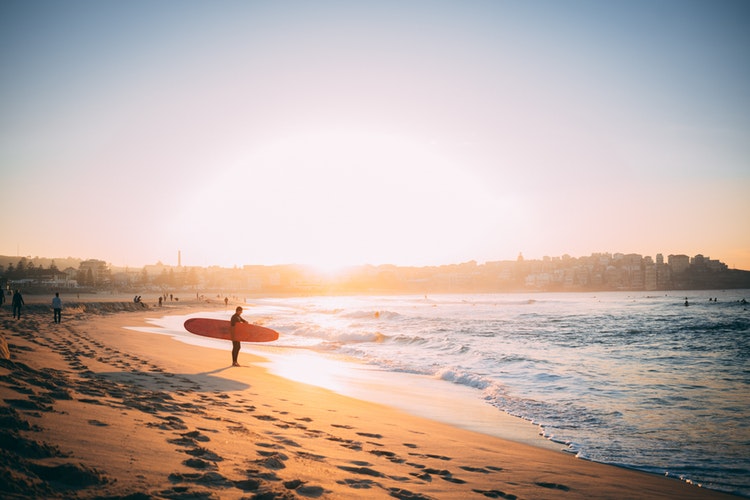 A male surfer stands on the shoreline of Bondi Beach with a surf board