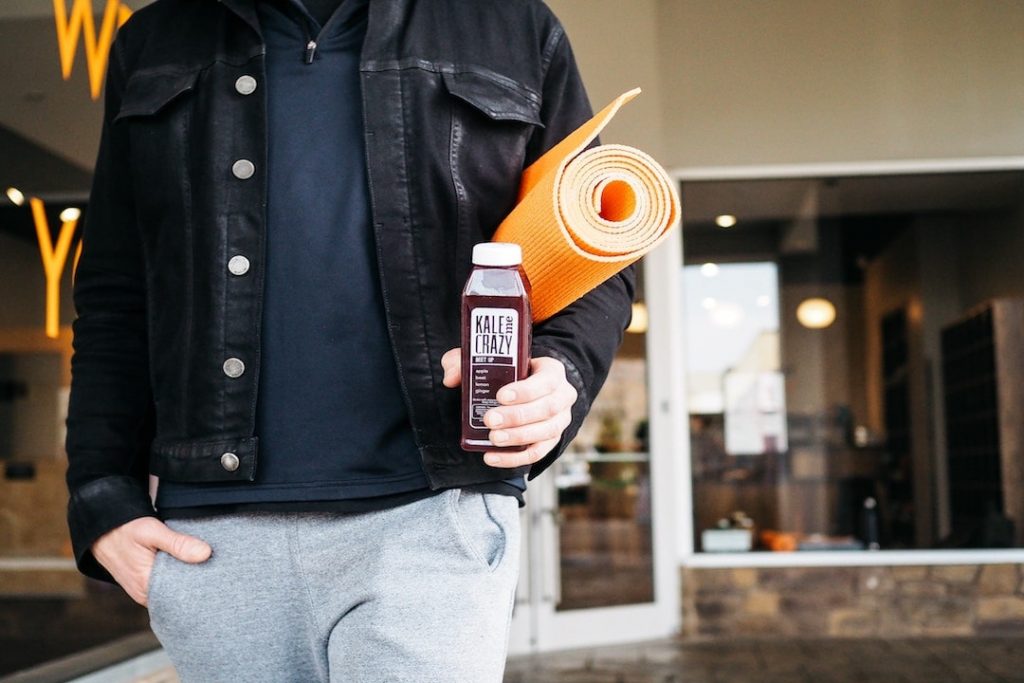 A person wearing a black jean jacket walks out of a building carrying an orange yoga mat