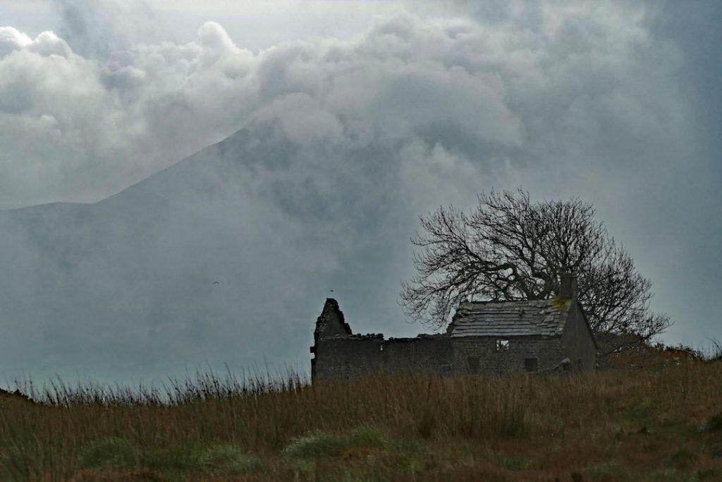 a ruin of an hold house in front of a leafless tree and a large hill on a foggy morning