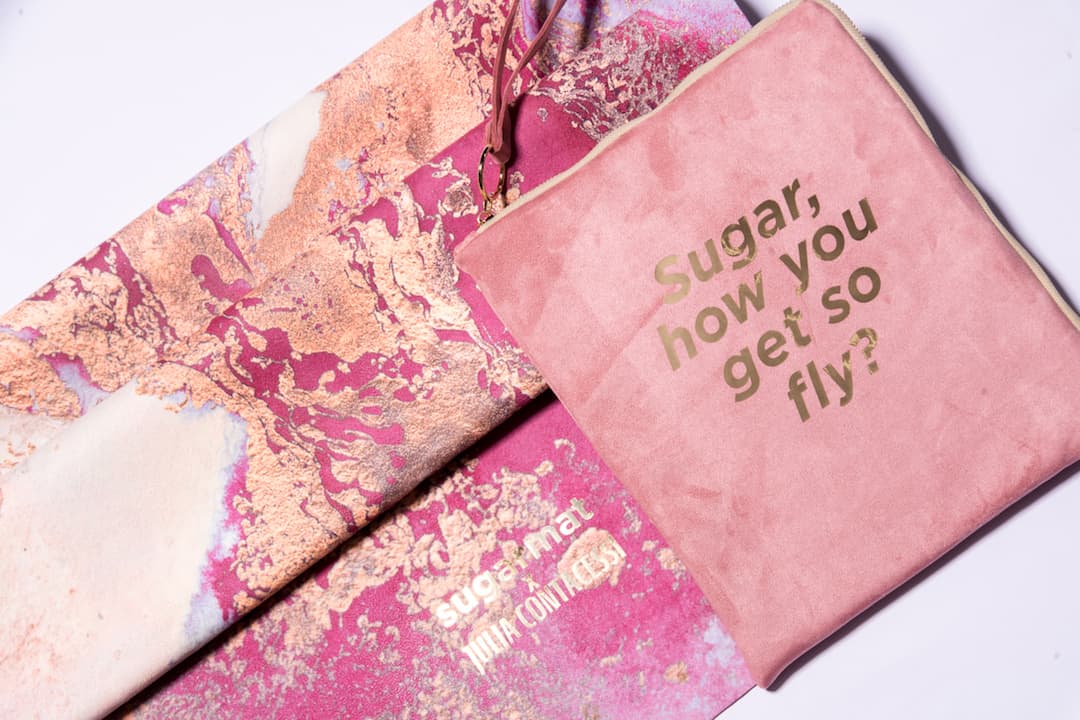 a close-up of a marbled pink yoga mat beside a travel pouch that reads "Sugar, how you get so fly?"