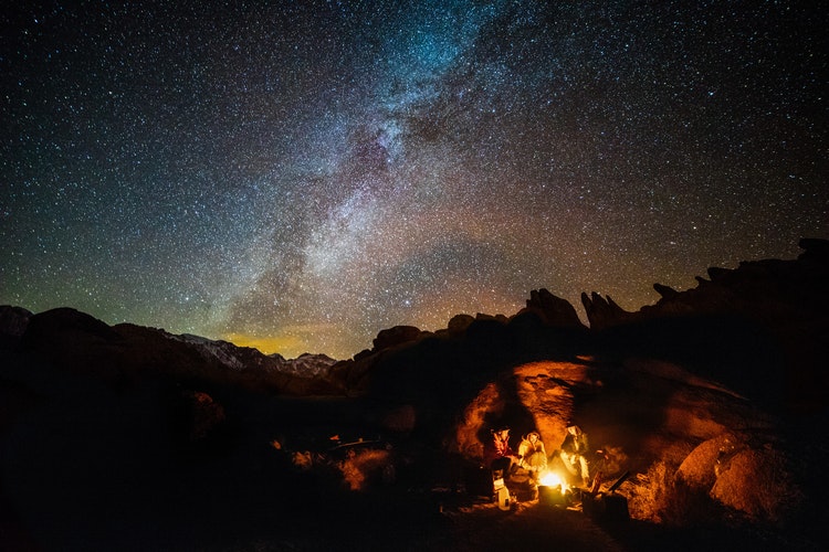 Safety Tips for Hiking at Night: a campfire burns below a starry sky around which people have gathered
