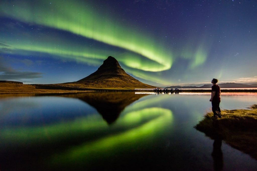 A man stands by a lake and stares up at the Northern Lights in Iceland
