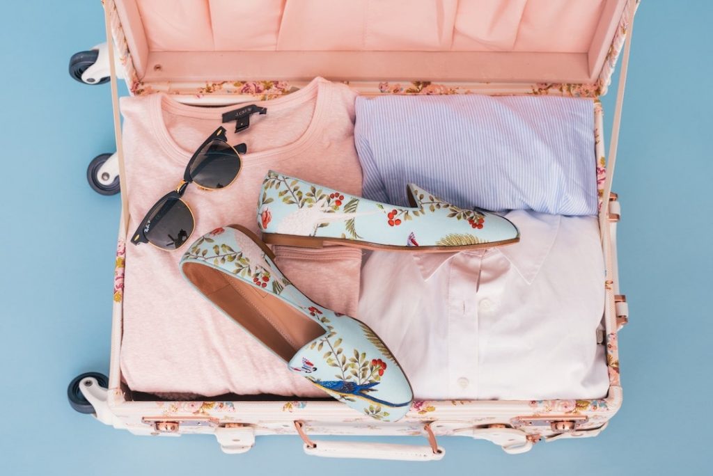 An open pink suitcase with pink and purple clothing, sunglasses, and shoes inside
