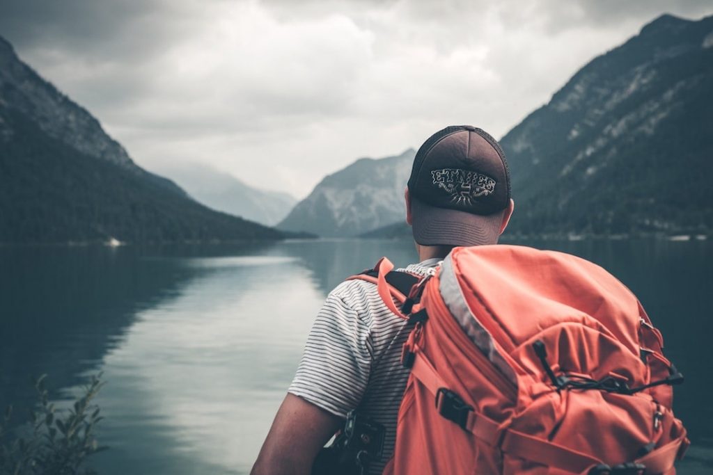 A person wearing an orange backpack stands in front of a lake at the base of mountains