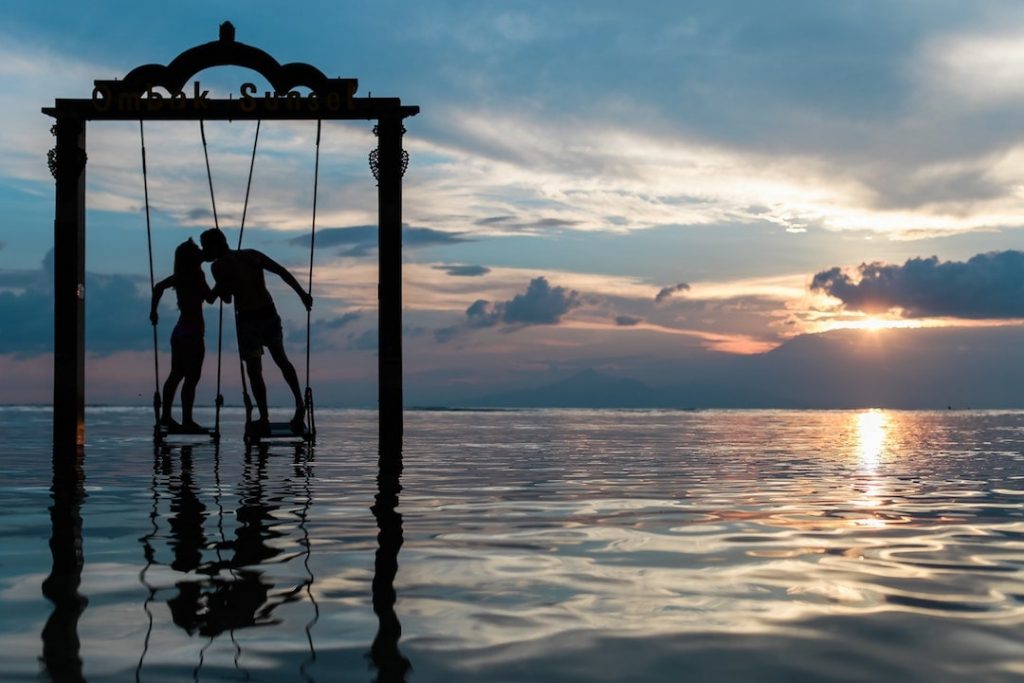 A couple on a swing over the ocean kiss at sunset