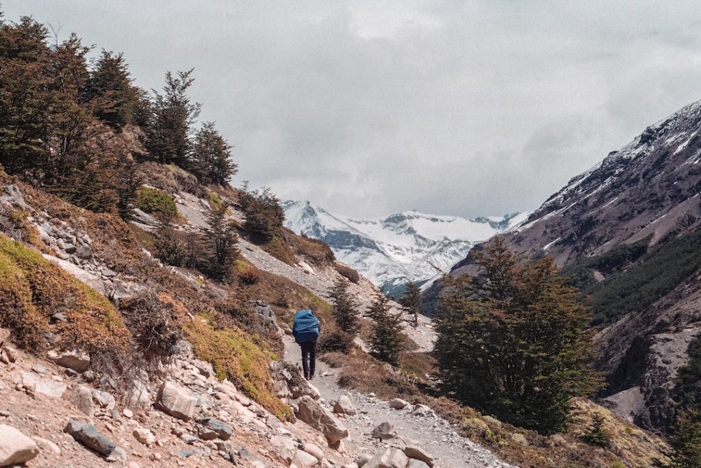 man in blue jacket hiking along a trail with snowcapped mountains in the background