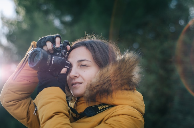 A woman in a yellow coat looks through the lens of a nikon camera