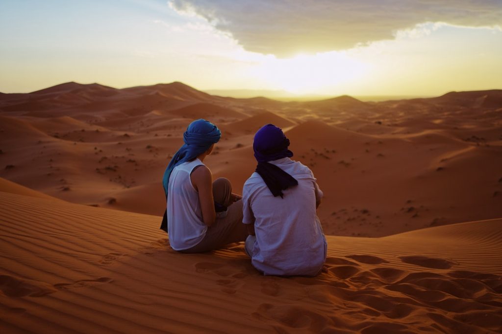 two people sitting on sand dunes with the sun in the background