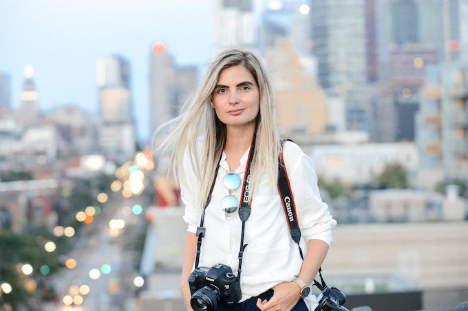 a woman in a white shirt with a camera around her neck with a city scape in blurred in the background
