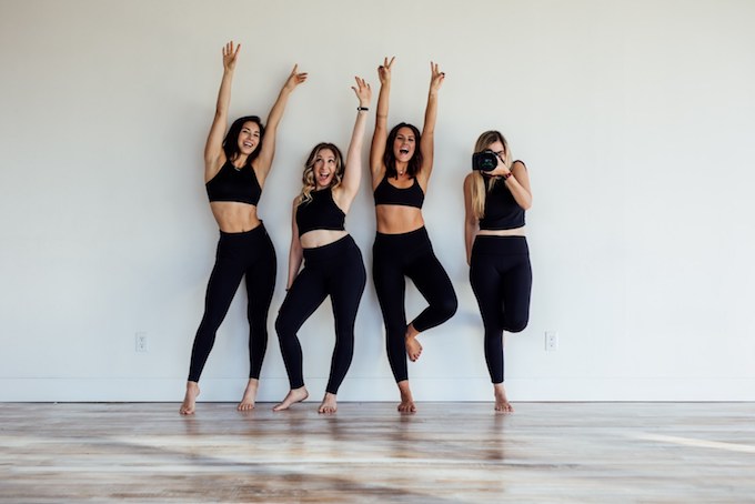 four women in a black leggings and crop tops stand against a white wall with their arms up in the air