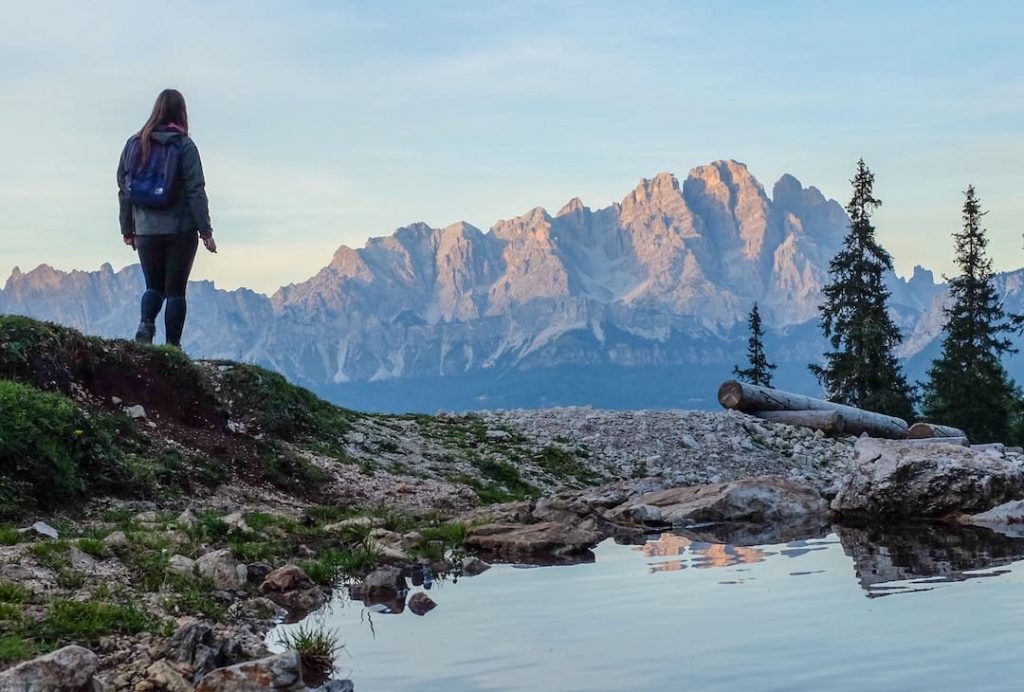 A woman standing beside a body of water and trees with mountains in the distance in Italy