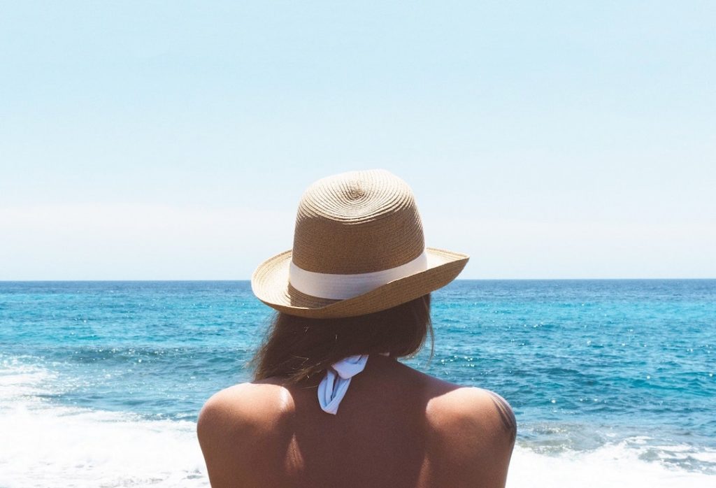 Woman standing on a beach facing the ocean wearing a hat