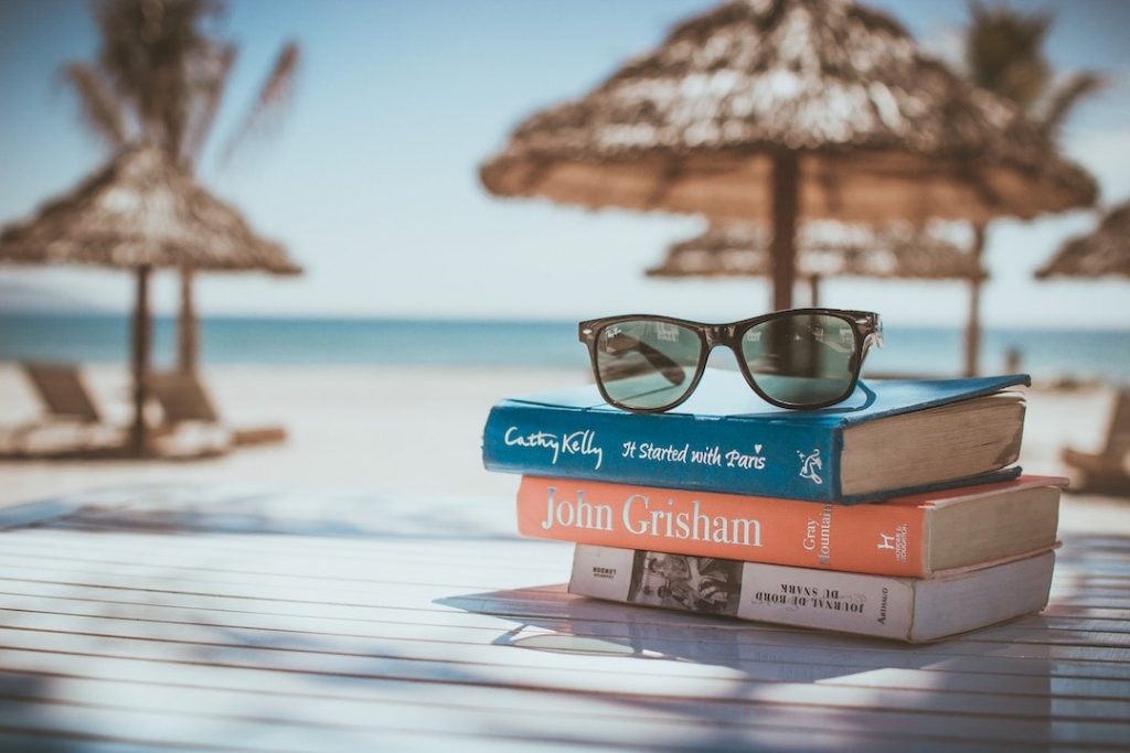 A pair of sunglasses sitting atop three books with a beach, lounge chairs, and umbrellas in the background