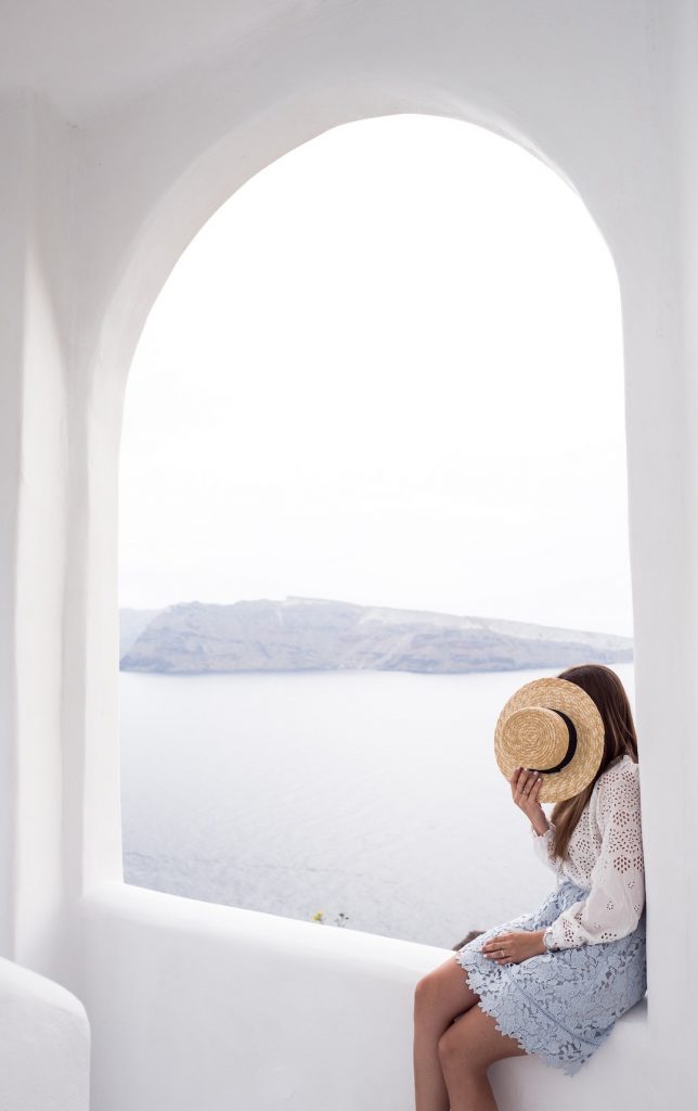 Woman posing with a hat in front of her face with Santorini scenery in the background