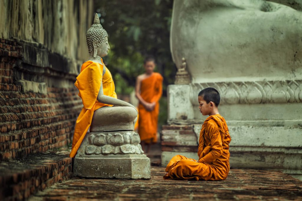 A young monk can be seen meditating in front of a Buddha statue in a temple in Thailand.