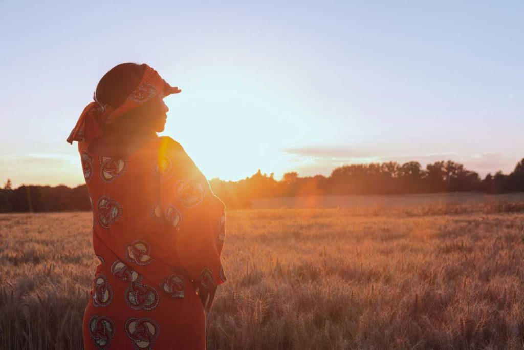 South African woman in traditional dress shades her eyes against the glare of the sun in a field of tall grass.