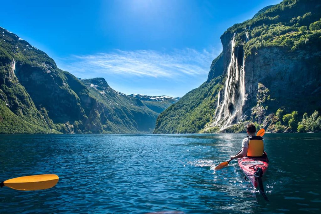 A male kayaker is paddling through the sparkling blue water of the Geiranger Fjord near the Seven Sisters Waterfall, Norway.