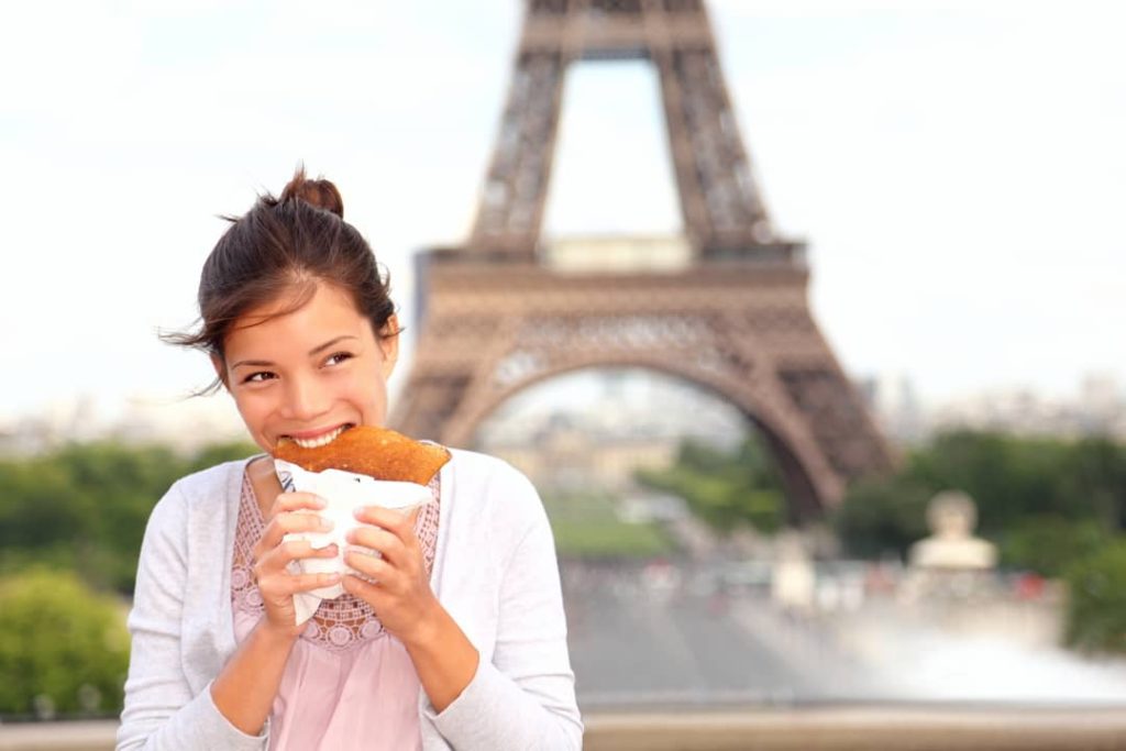 Woman bites into a tasty crepe as she stands with her back to the beautiful Eiffel Tower in Paris, France.